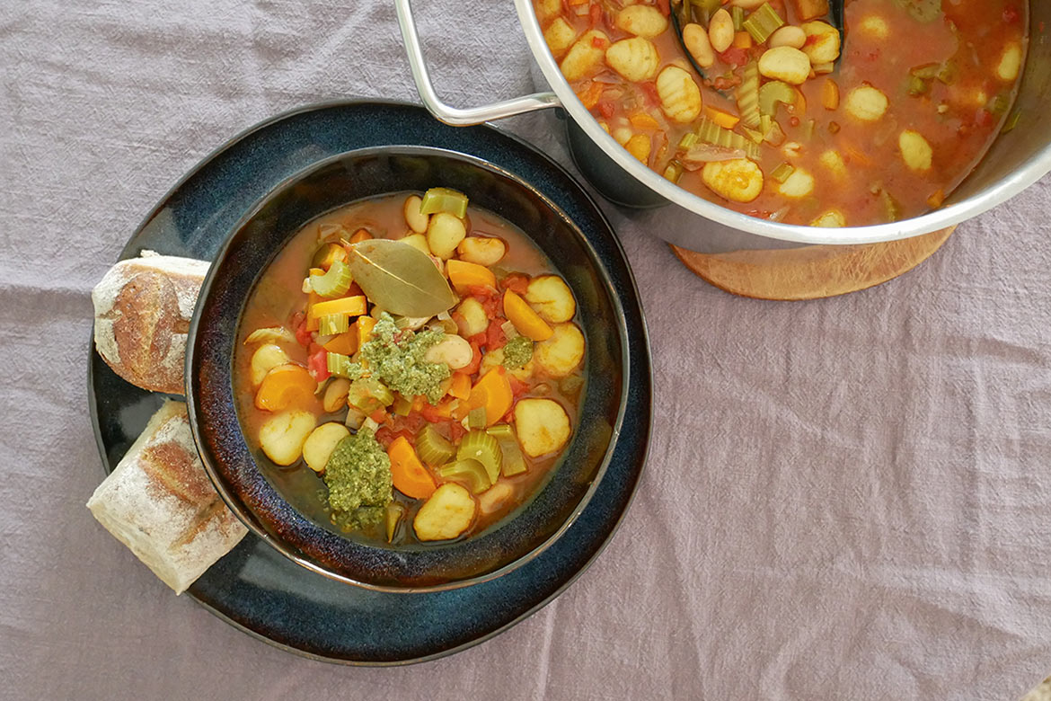 Auf einem Tisch steht eine dampfende Schüssel Minestrone und ein Topf mit der italienischen Suppe.