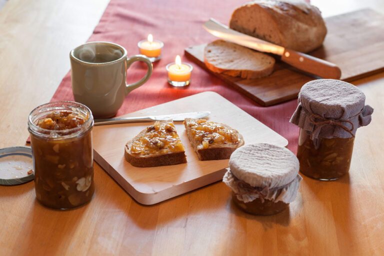 Drei Gläser mit Bratapfelmarmelade stehen auf dem Frühstückstisch, eine Scheibe Brot ist mit der Marmelade bestrichen.