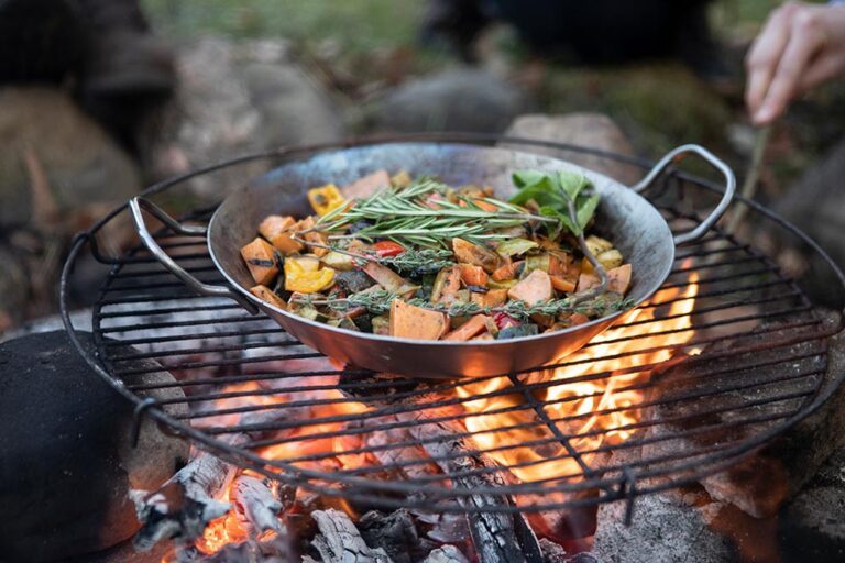 Über einem Lagerfeuer wird beim Outdoor-Kochen Gemüse in einer Eisenpfanne gegrillt.