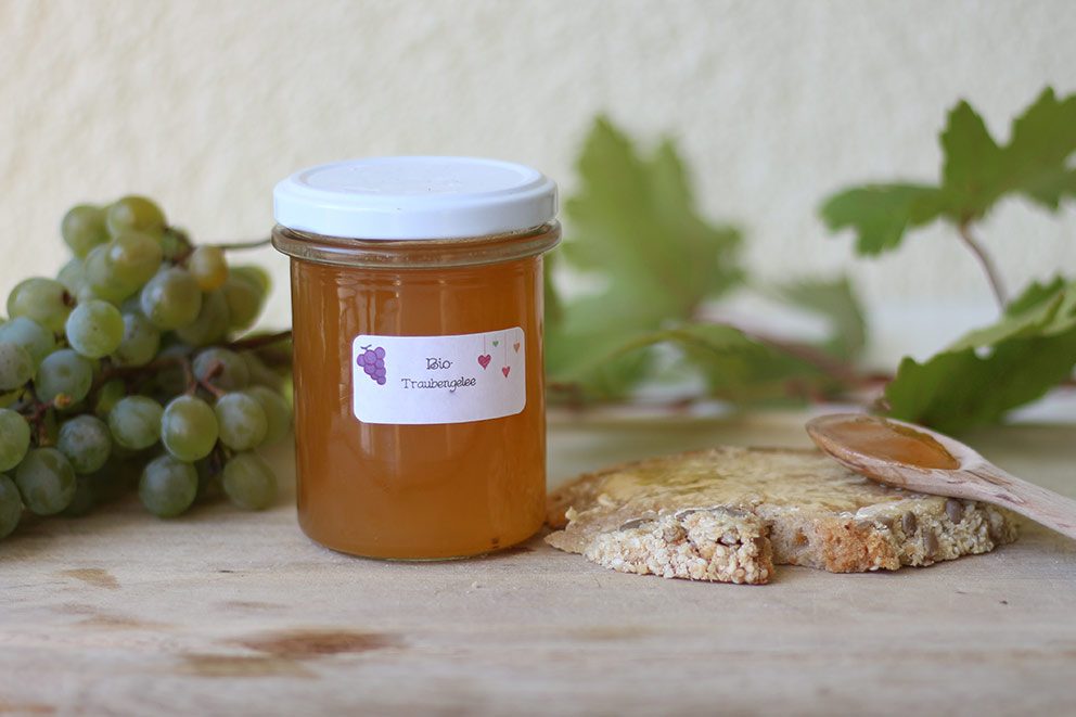 Ein Glas mit selbst gemachtem Traubengelee steht auf einem Holzbrettchen neben einer Scheibe Brot.