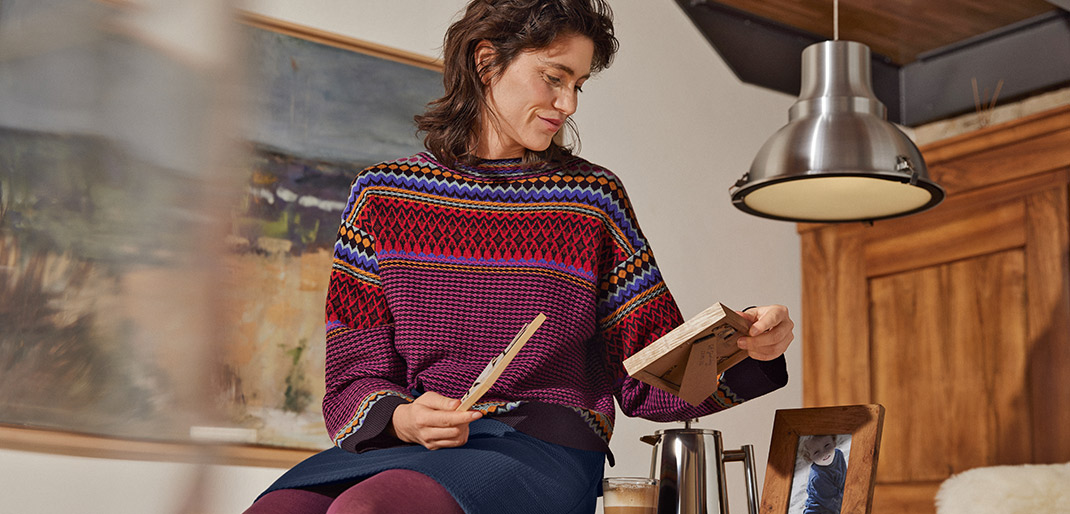 Frau trägt einen Jacquard-Strickpullover, sitzt auf einem Tisch und betrachtet ein Bild.