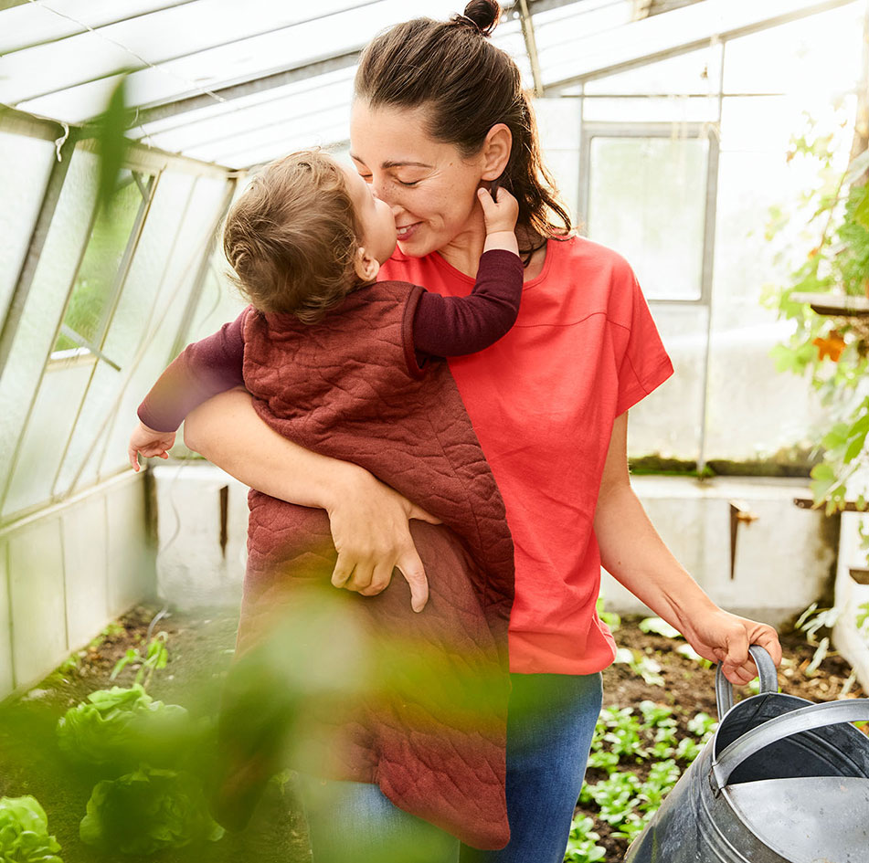 Baby in Stepp-Schlafsack auf dem Arm seiner Mutter in einem Gewächshaus